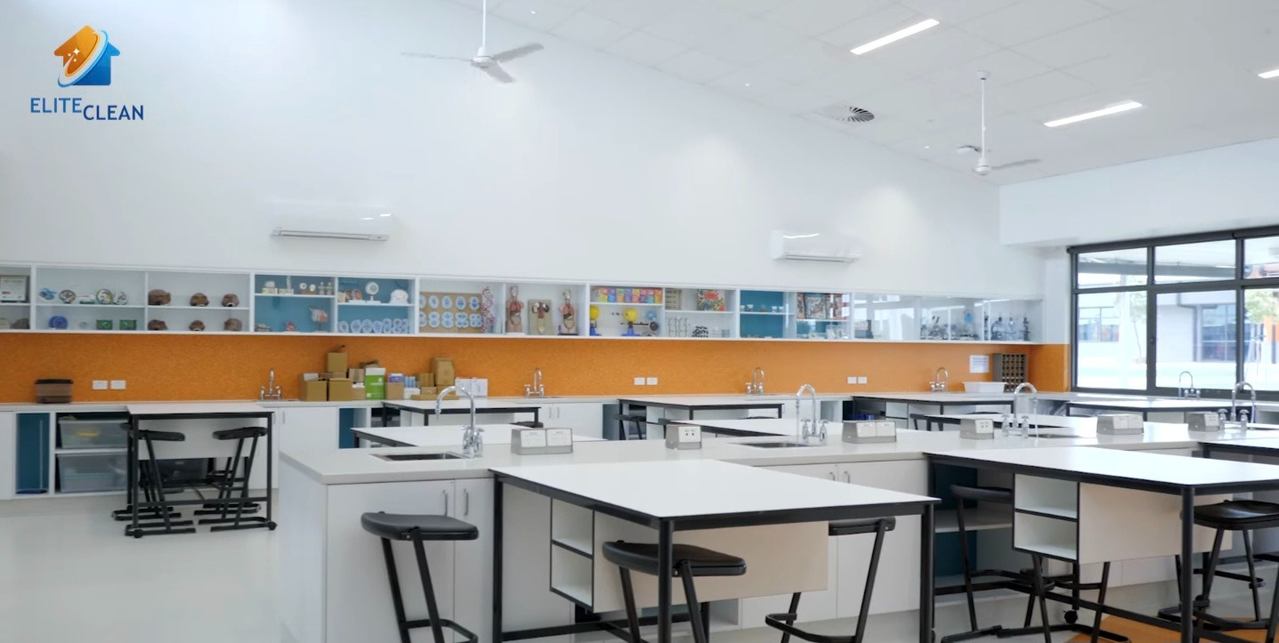 Wnętrze czystej, jasnej pracowni chemicznej z białymi blatami, czarnymi stołkami i pomarańczową mozaiką. Na ścianie logo firmy sprzątającej.