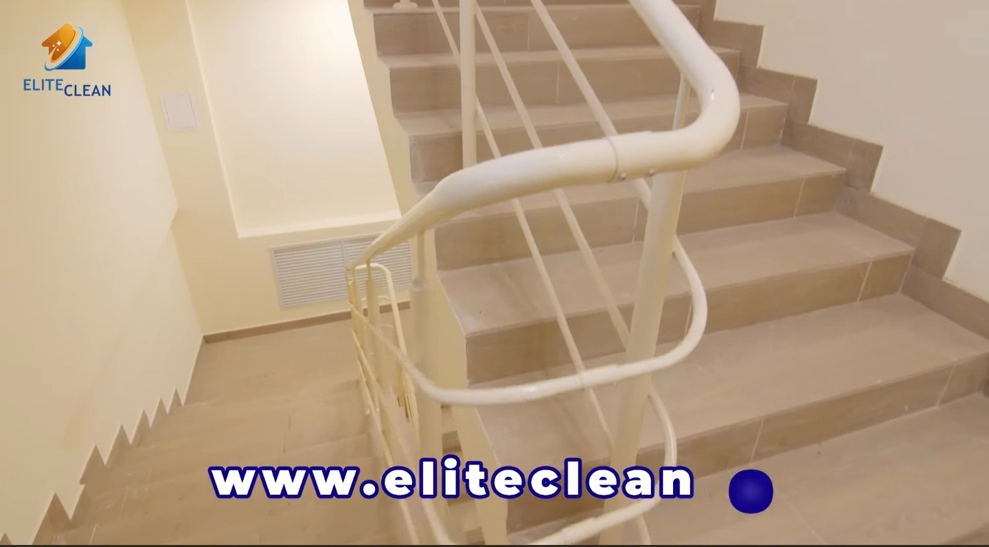 Klatka schodowa z beżowymi płytkami i białą balustradą, widok z góry. Czyste schody i ściany. Na dole widoczny adres strony internetowej firmy sprzątającej.