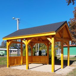 WoodWizard Mariusz Jarosik - Żółta, drewniana konstrukcja z czarnym dachem na betonowej podstawie, widok z zewnątrz w słoneczny dzień.