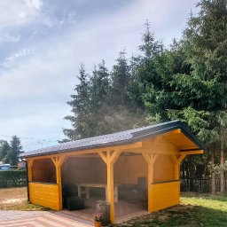 WoodWizard Mariusz Jarosik - Żółta, drewniana konstrukcja z czarnym dachem, prawdopodobnie altana, stoi na trawniku obok wzorzystego chodnika z kostki, w tle wysokie drzewa iglaste.