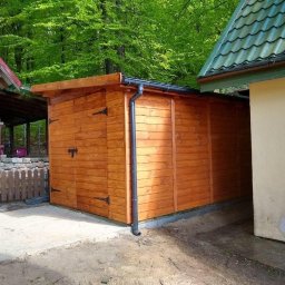 WoodWizard Mariusz Jarosik - Drewniana konstrukcja z czarnymi okuciami i rynną, przylegająca do ściany budynku, na tle zieleni drzew