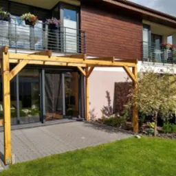 Drewniana konstrukcja z belek nad szarym tarasem z kostki brukowej, przylegająca do nowoczesnego domu z balkonami i kwiatami w donicach.