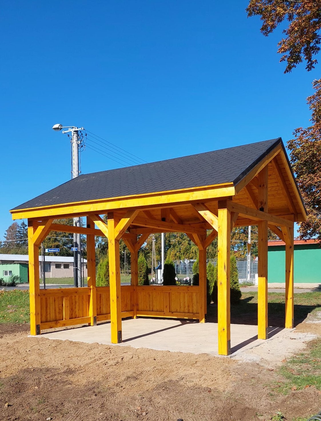 Żółta, drewniana konstrukcja z czarnym dachem na betonowej podstawie, widok z zewnątrz w słoneczny dzień.