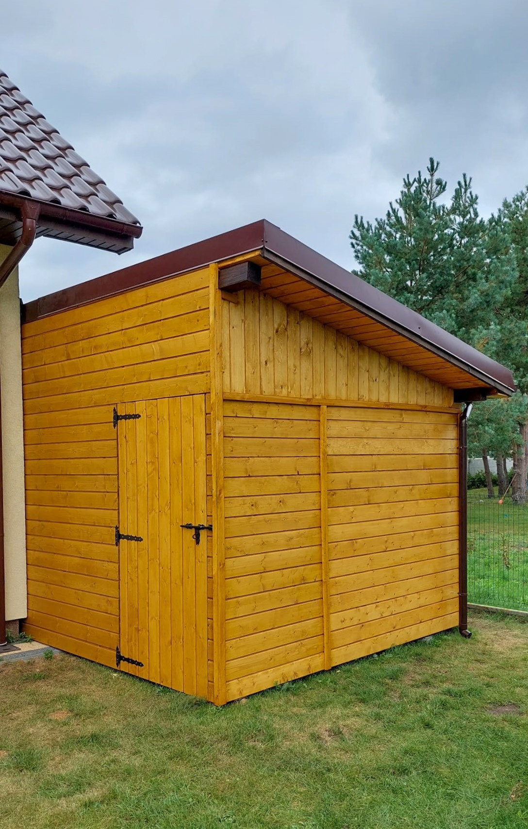 Złocista, drewniana konstrukcja z drzwiami i brązowym dachem na trawniku, przylegająca do budynku z brązowym dachem i rynną, na tle drzew i pochmurnego nieba.