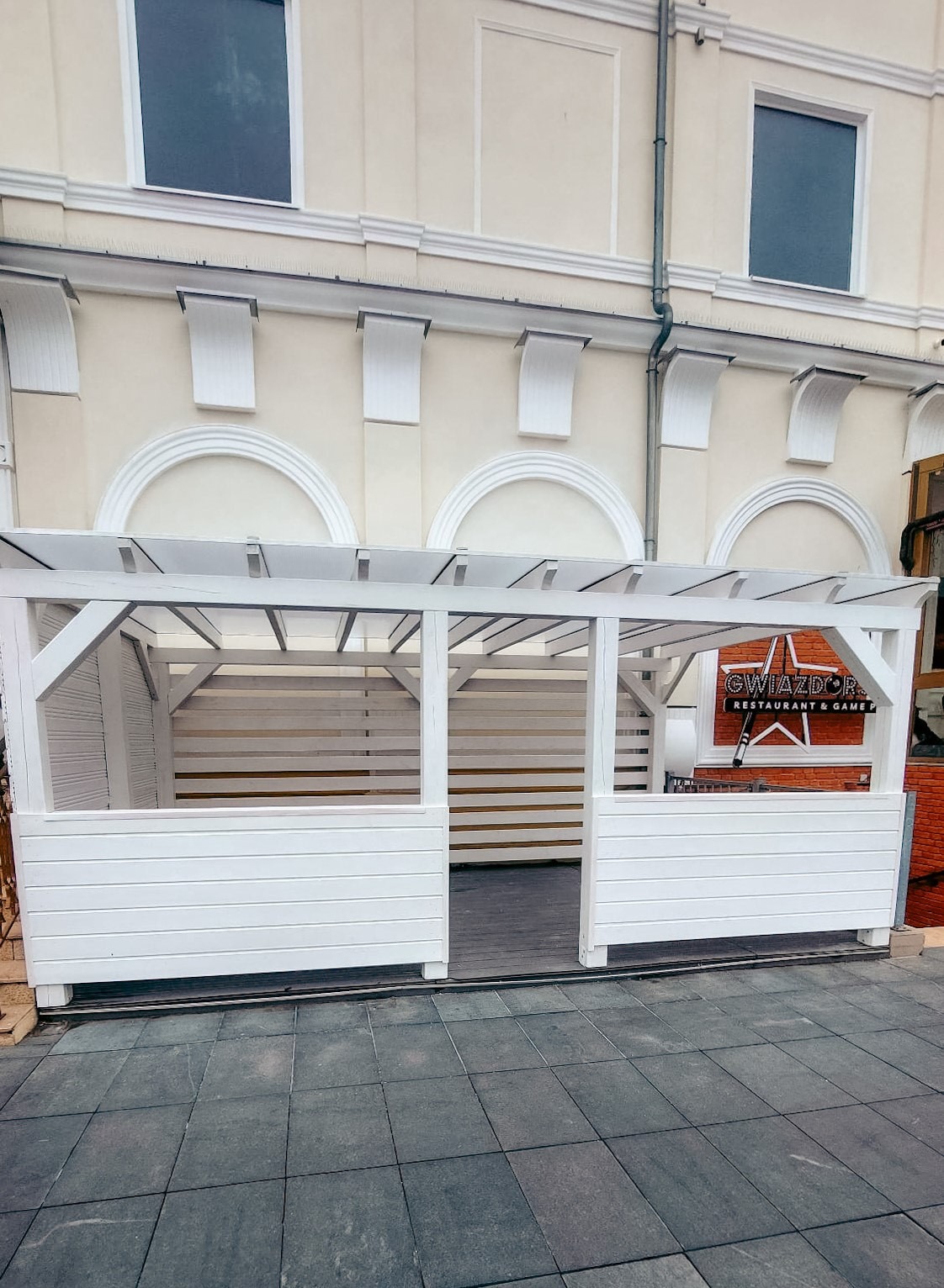 Biała, drewniana konstrukcja z ażurowymi ścianami, prawdopodobnie zewnętrzna strefa restauracji z widocznym logo 'Gwiazdor', umieszczona na szarym bruku.