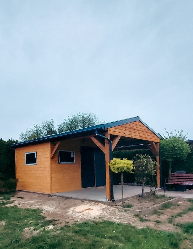 Drewniana konstrukcja z zadaszeniem, przypominająca małą altanę z dobudowanym pomieszczeniem gospodarczym, wykończona w naturalnym odcieniu drewna, z ciemnymi elementami dachu i drzwi, na tle...