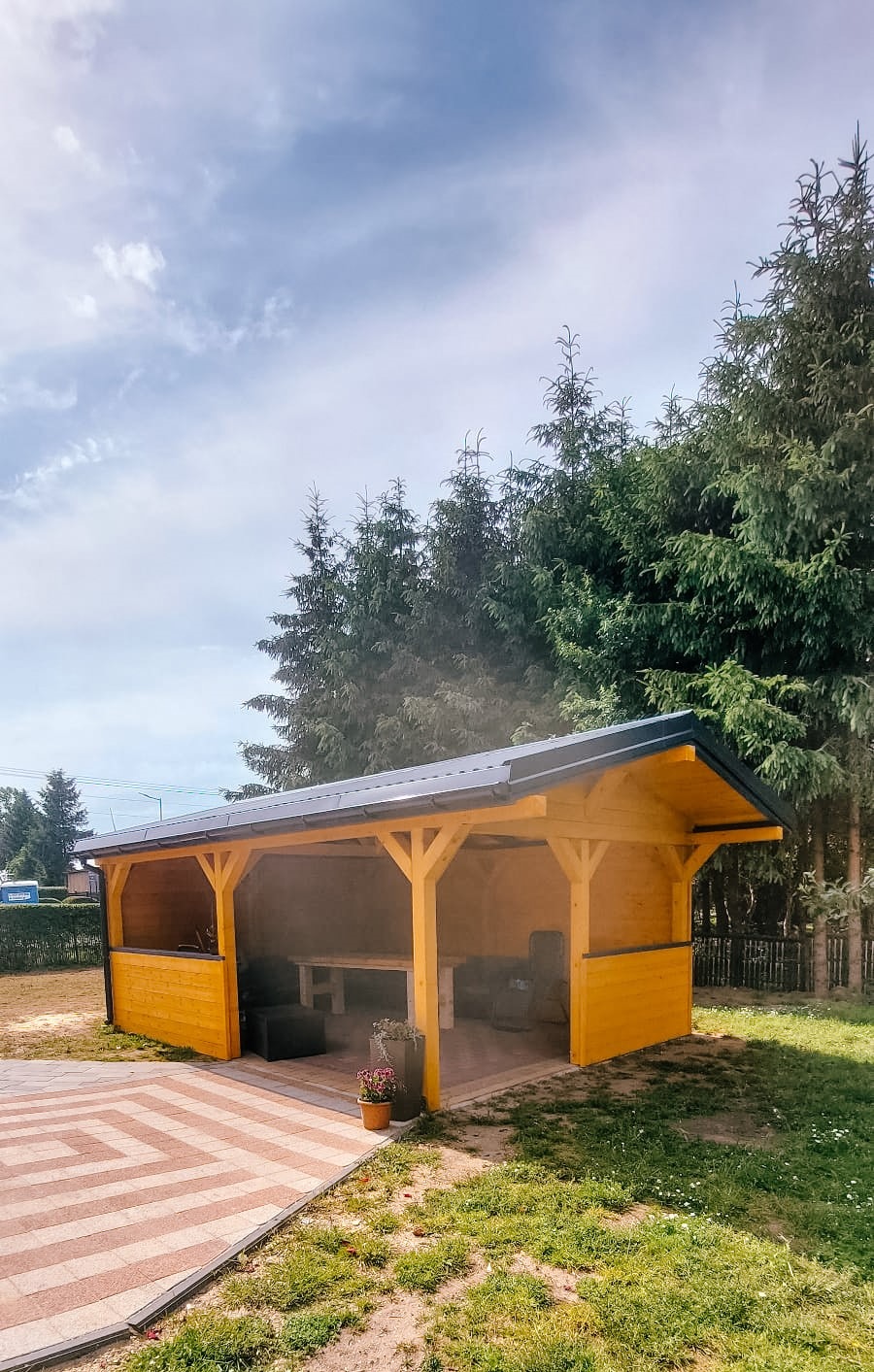 Żółta, drewniana konstrukcja z czarnym dachem, prawdopodobnie altana, stoi na trawniku obok wzorzystego chodnika z kostki, w tle wysokie drzewa iglaste.