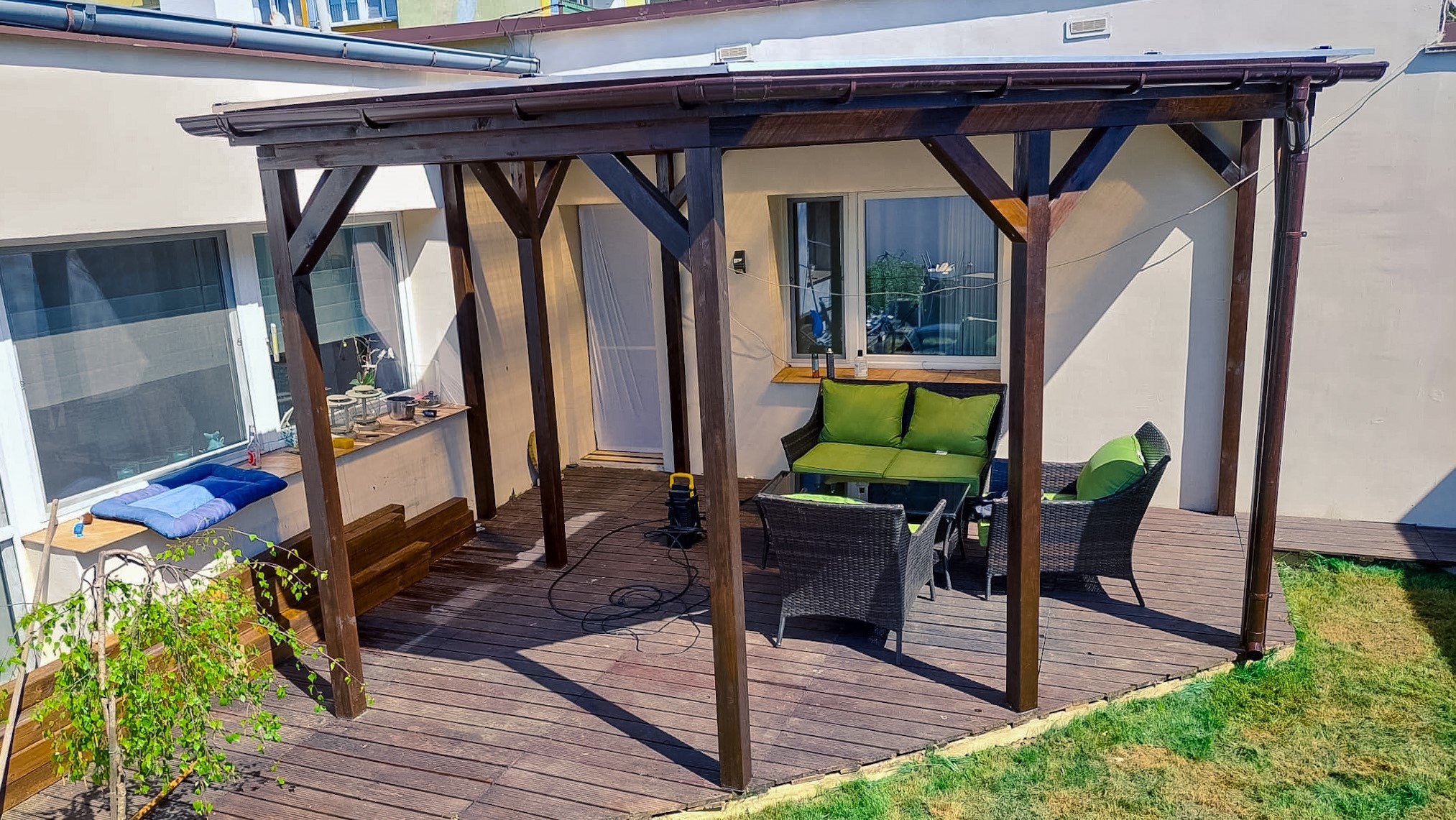 Ciemnobrązowa pergola drewniana z meblami ogrodowymi i tarasem z desek, widok z zewnątrz, obok domu z białymi ścianami i zielonym trawnikiem.