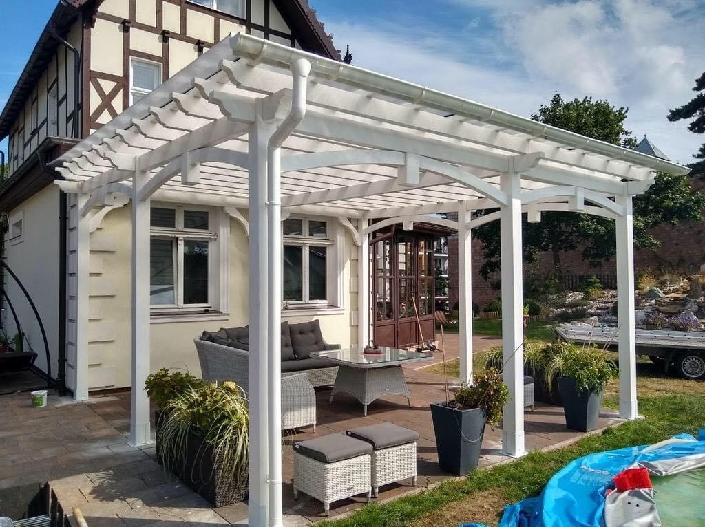 Biała konstrukcja z drewna nad tarasem przylegającym do budynku z oknami, meble ogrodowe z szarego rattanu, donice z roślinami, w tle ogród i przyczepka.