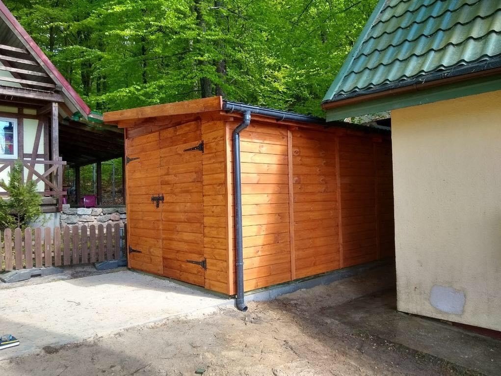 Drewniana konstrukcja z czarnymi okuciami i rynną, przylegająca do ściany budynku, na tle zieleni drzew