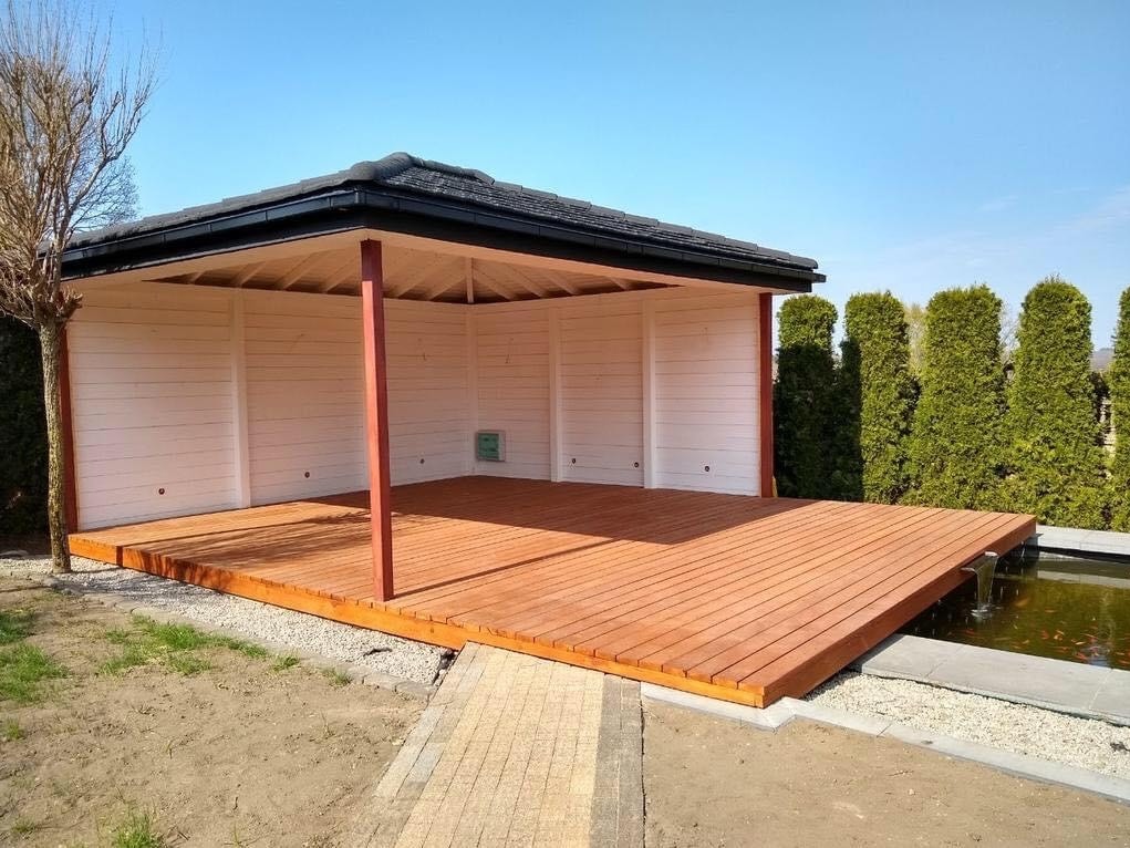 Otwarta, drewniana konstrukcja z płaskim dachem i drewnianą podłogą, częściowo otoczona białymi ścianami, przylegająca do oczka wodnego z kaskadą, widoczna z perspektywy ogrodu.