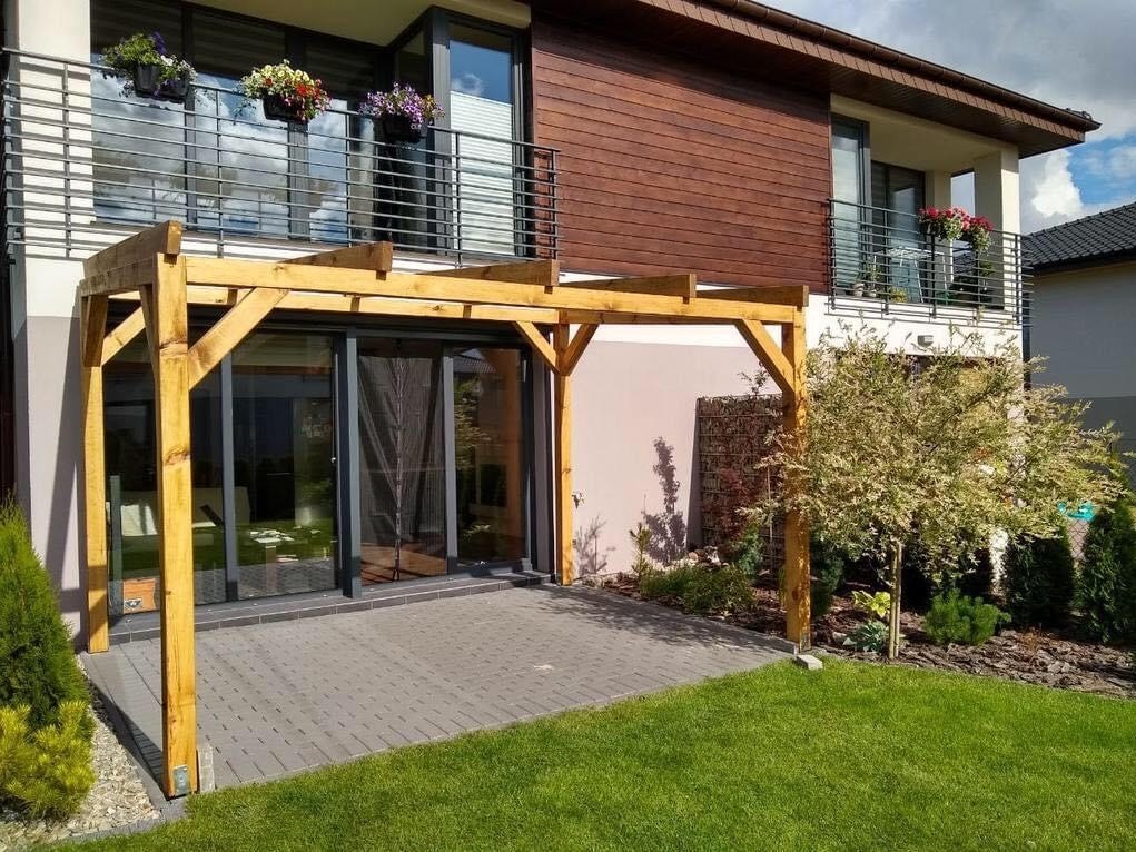 Drewniana konstrukcja z belek nad szarym tarasem z kostki brukowej, przylegająca do nowoczesnego domu z balkonami i kwiatami w donicach.