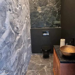 KAMA - Nowoczesna łazienka z dekoracyjną ścianą z efektem kamienia, szarą ścianą z fakturą, czarną toaletą i złotą umywalką na drewnianej szafce.