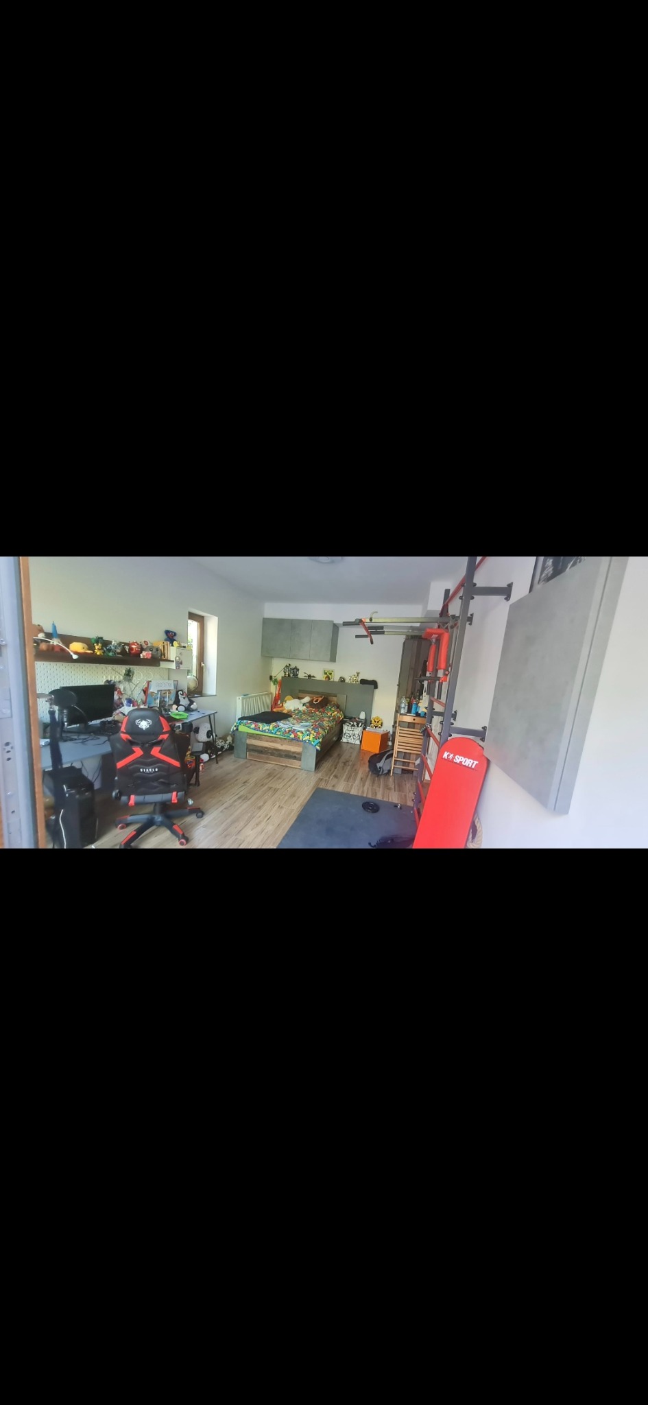 Pokój młodzieżowy z drewnianą podłogą, biurkiem z krzesłem gamingowym, łóżkiem z kolorową pościelą i ścianką do ćwiczeń z ławką do wyciskania.