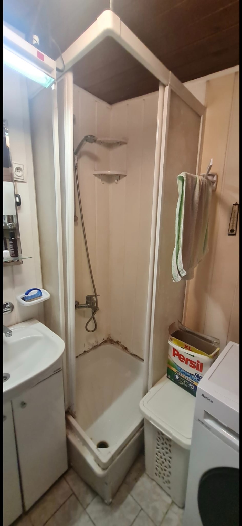 Narożna kabina prysznicowa z widocznymi śladami użytkowania i pleśnią w rogu, obok pralka i pojemnik na proszek do prania