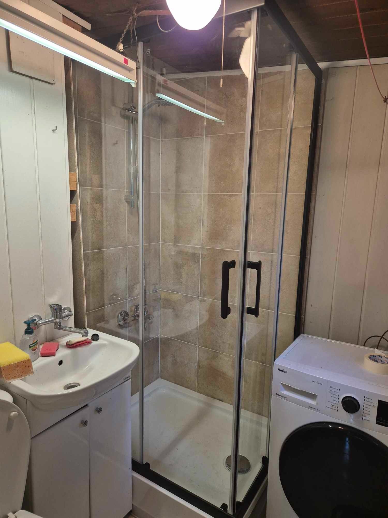 Kabina prysznicowa z czarnymi profilami i umywalka z szafką w małej łazience, obok pralka Amica; płytki w odcieniach beżu, biała armatura, sufit z widocznymi instalacjami.
