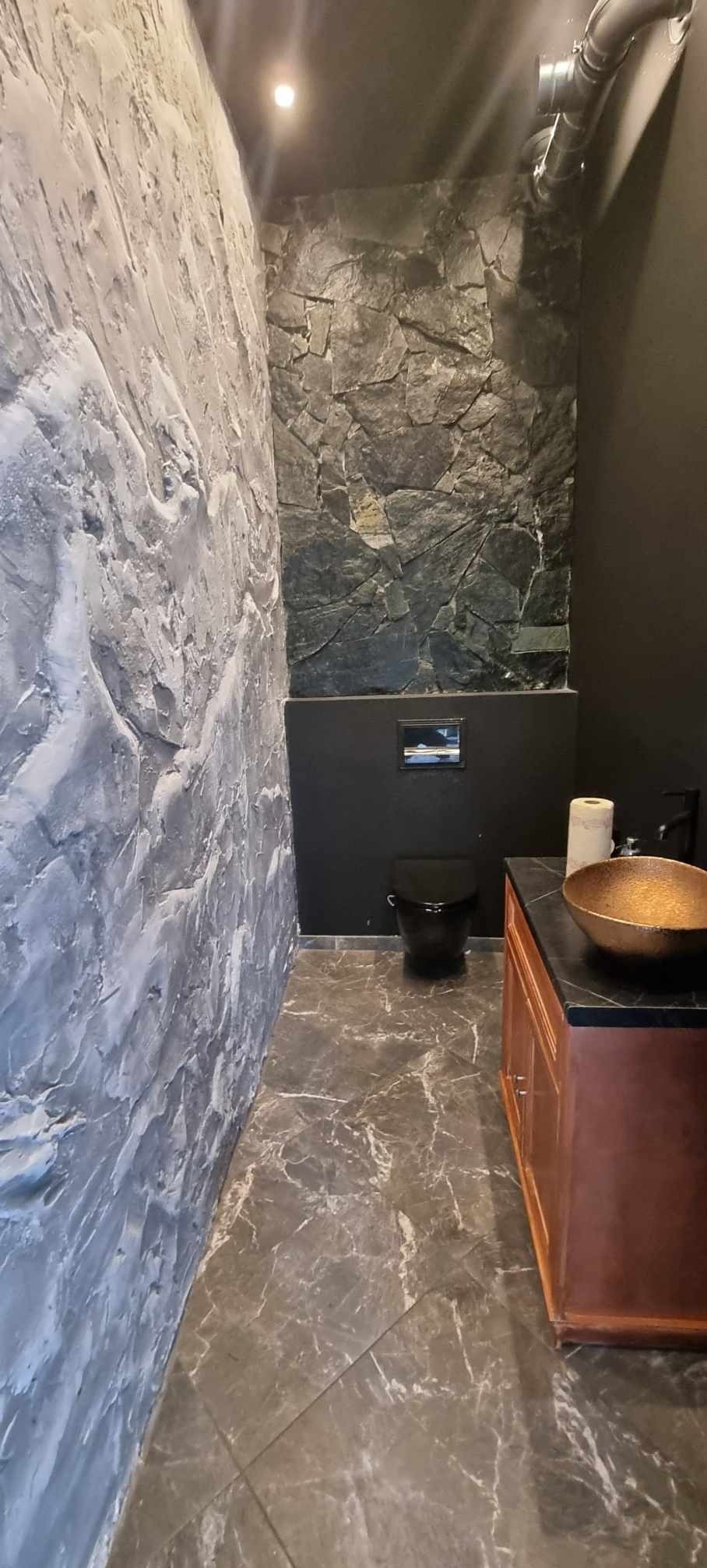 Nowoczesna łazienka z dekoracyjną ścianą z efektem kamienia, szarą ścianą z fakturą, czarną toaletą i złotą umywalką na drewnianej szafce.