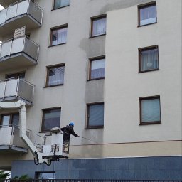RKM Mycie Elewacji - Pracownik w niebieskim kasku na podnośniku czyści elewację budynku. Widoczny efekt przed i po myciu. Szara elewacja, okna i balkony.