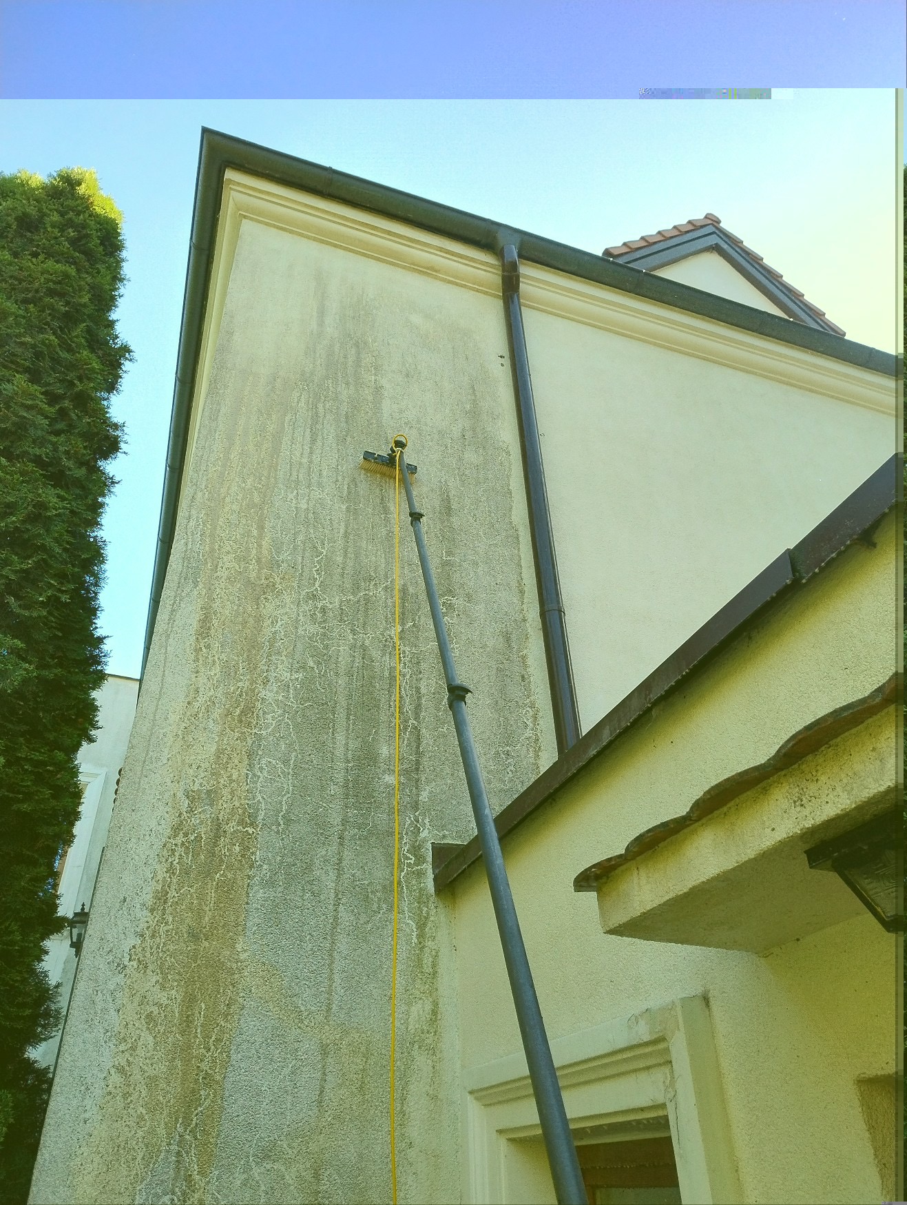 Czyszczenie elewacji budynku z użyciem teleskopowej lancy i szczotki. Widoczny fragment zabrudzonej ściany i fragment czystej elewacji. Praca w toku.
