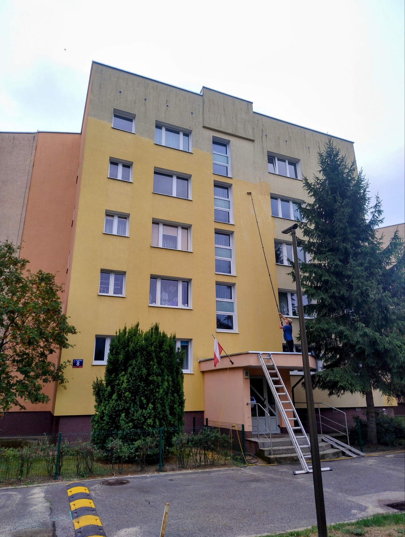Pracownik na drabinie myje żółtą elewację bloku za pomocą długiej lancy. Część ściany jest już umyta, ukazując jaśniejszy odcień. Widoczny polski flaga.