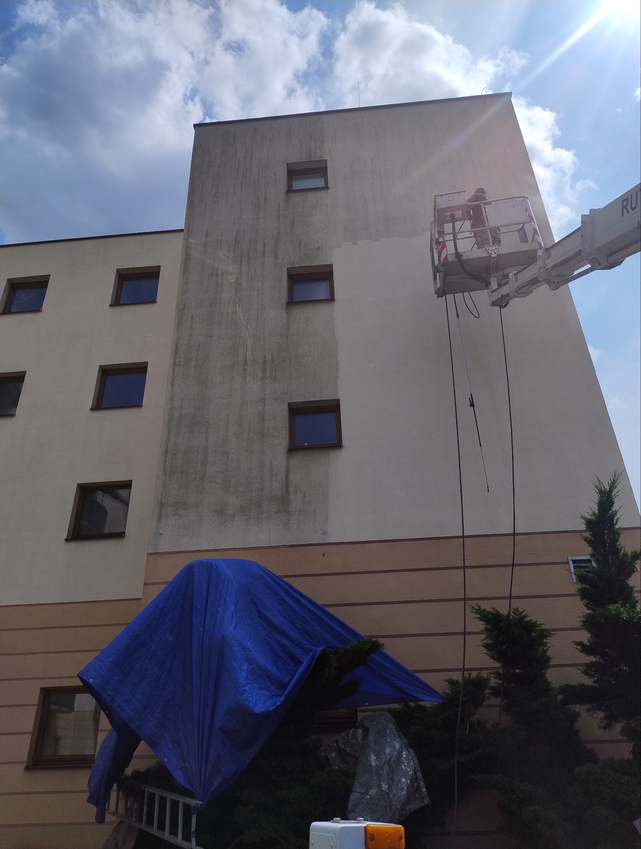 Mycie elewacji budynku z podnośnika koszowego. Widoczna różnica między umytą a brudną częścią ściany. Na dole zabezpieczone rośliny plandeką.