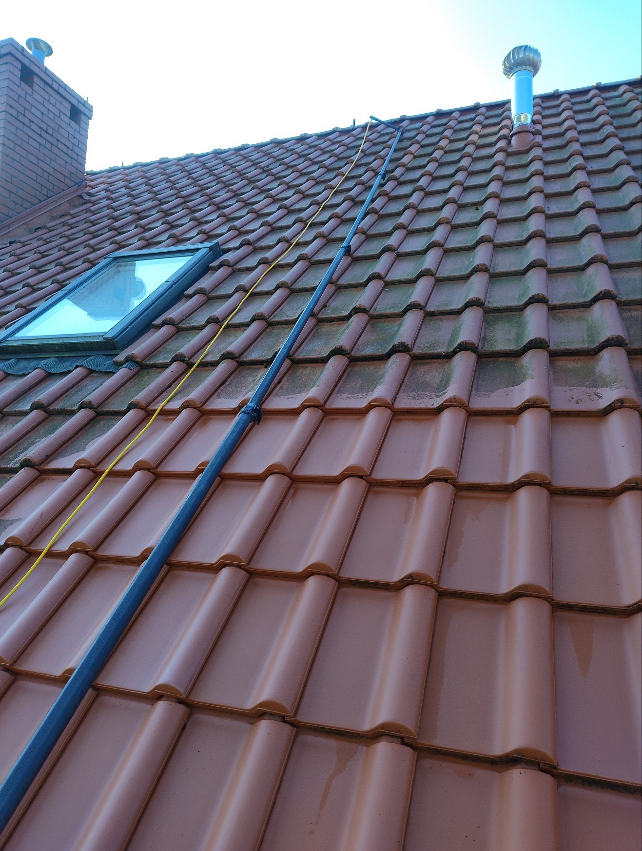Czerwona dachówka po czyszczeniu w Warszawie. Widoczna lanca teleskopowa i okno dachowe. Część dachu jest umyta, a część jeszcze nie. Kominek.
