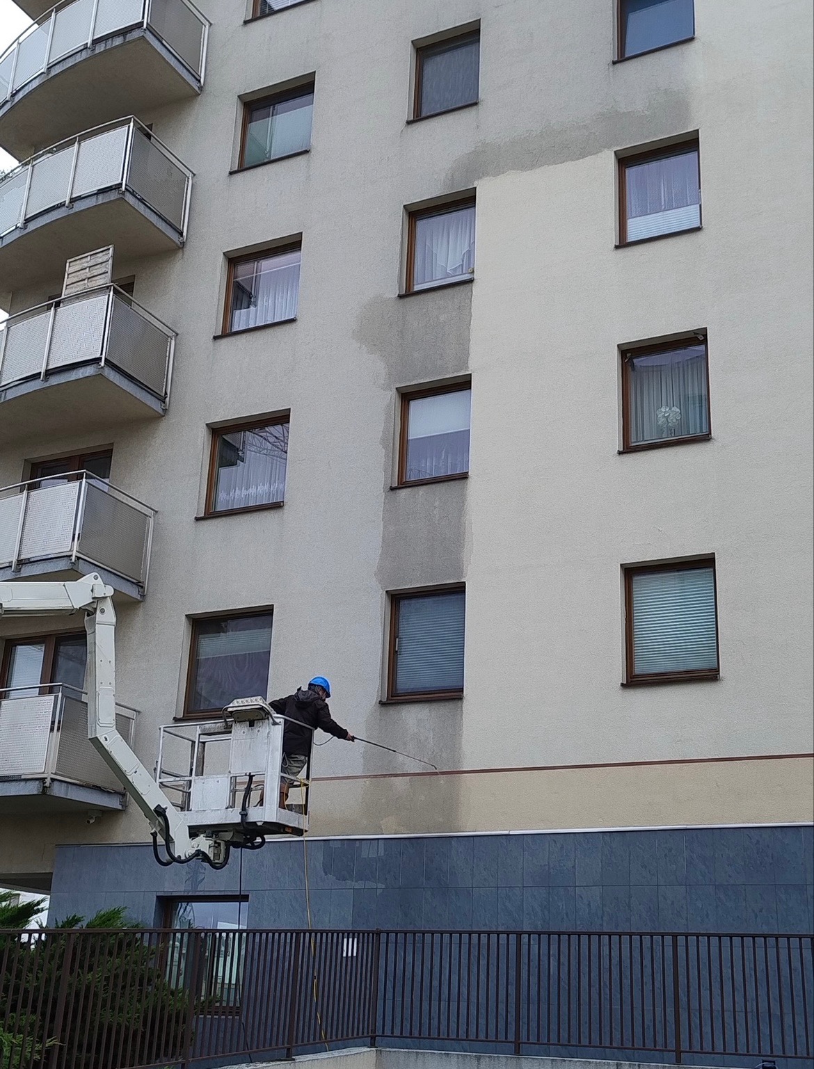 Pracownik w niebieskim kasku na podnośniku czyści elewację budynku. Widoczny efekt przed i po myciu. Szara elewacja, okna i balkony.