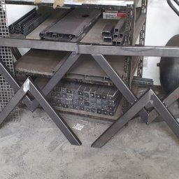 VTECH - Elementy konstrukcji stalowej w warsztacie: spawana rama stołu z profili, na tle regałów z metalowymi elementami i kompresora.