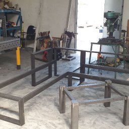 VTECH - Warsztatowa przestrzeń z metalowymi konstrukcjami w trakcie spawania, widoczny stół spawalniczy z otworami, w tle wiertarka stołowa i narzędzia.