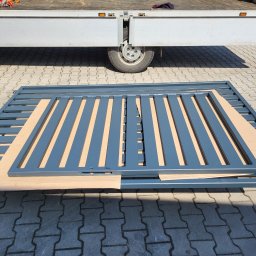 VTECH - Pomazana na szaro metalowa brama wjazdowa, zabezpieczona kartonem podczas transportu, leży na kostce brukowej, w tle widoczny fragment samochodu dostawczego.