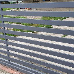 VTECH - Nowoczesna, pozioma brama wjazdowa z szarych, metalowych profili, widziana z bliska, z kostką brukową u dołu i zielenią w tle.