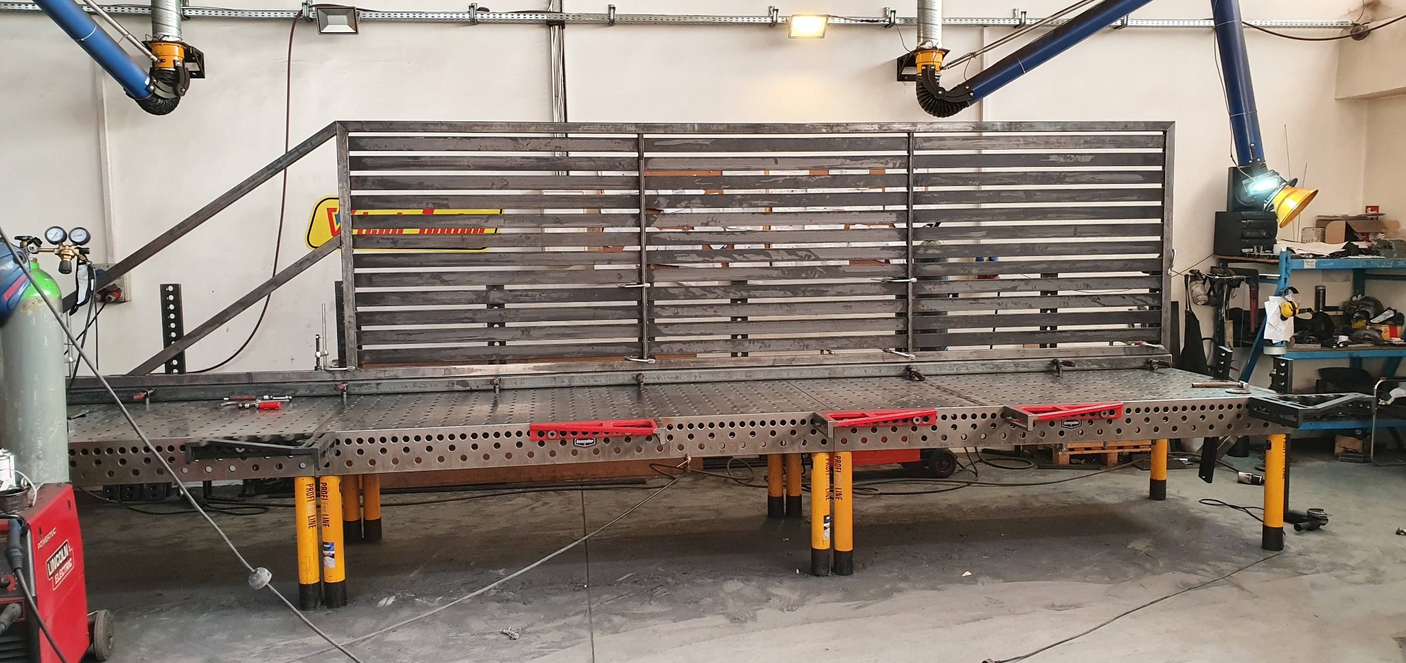 Spawanie metalowej konstrukcji z poziomych profili na perforowanym stole spawalniczym z żółtymi nogami, widoczne narzędzia i sprzęt spawalniczy.