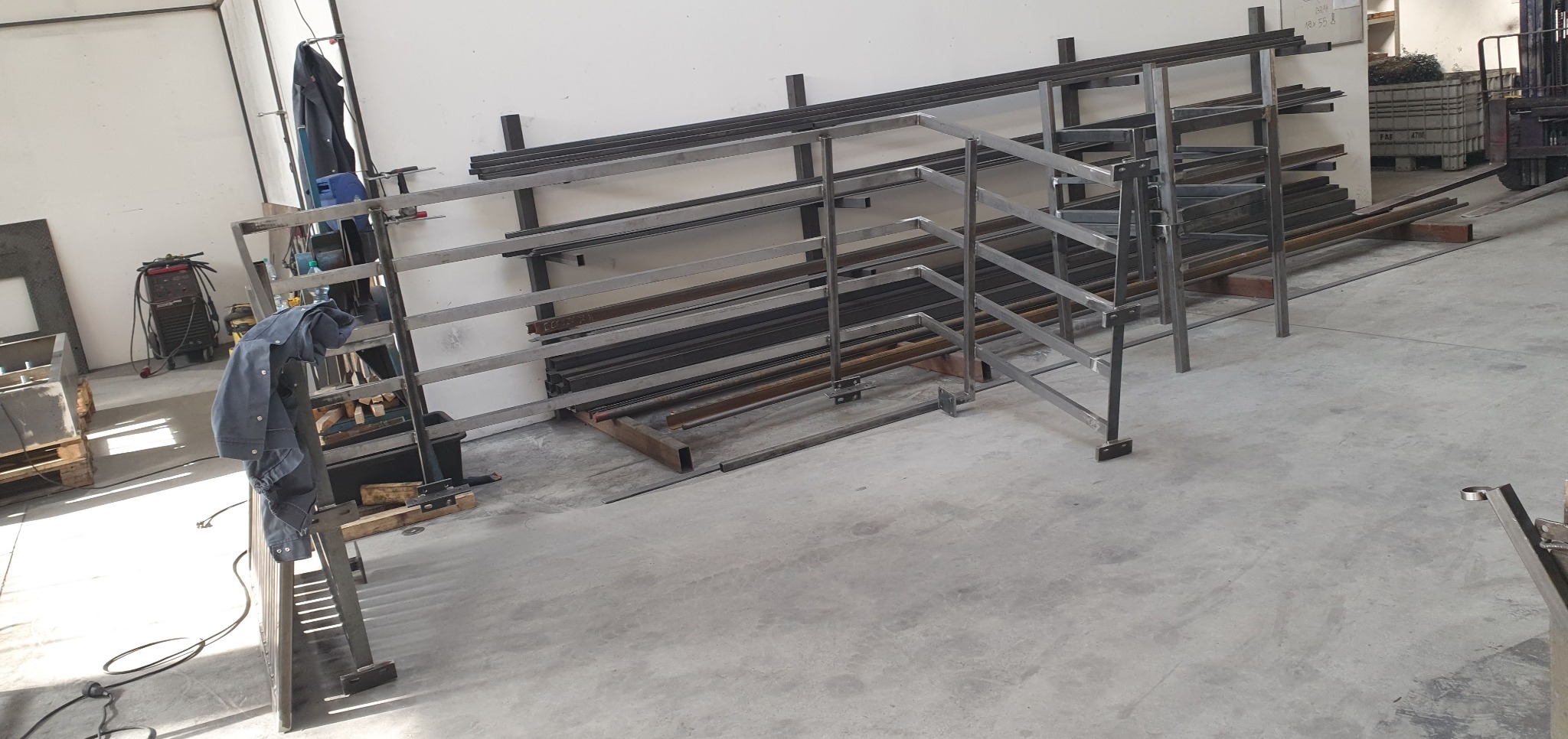 Wnętrze warsztatu z metalowymi konstrukcjami regałowymi i elementami poręczy w trakcie produkcji, widoczny kombinezon roboczy i spawarka.