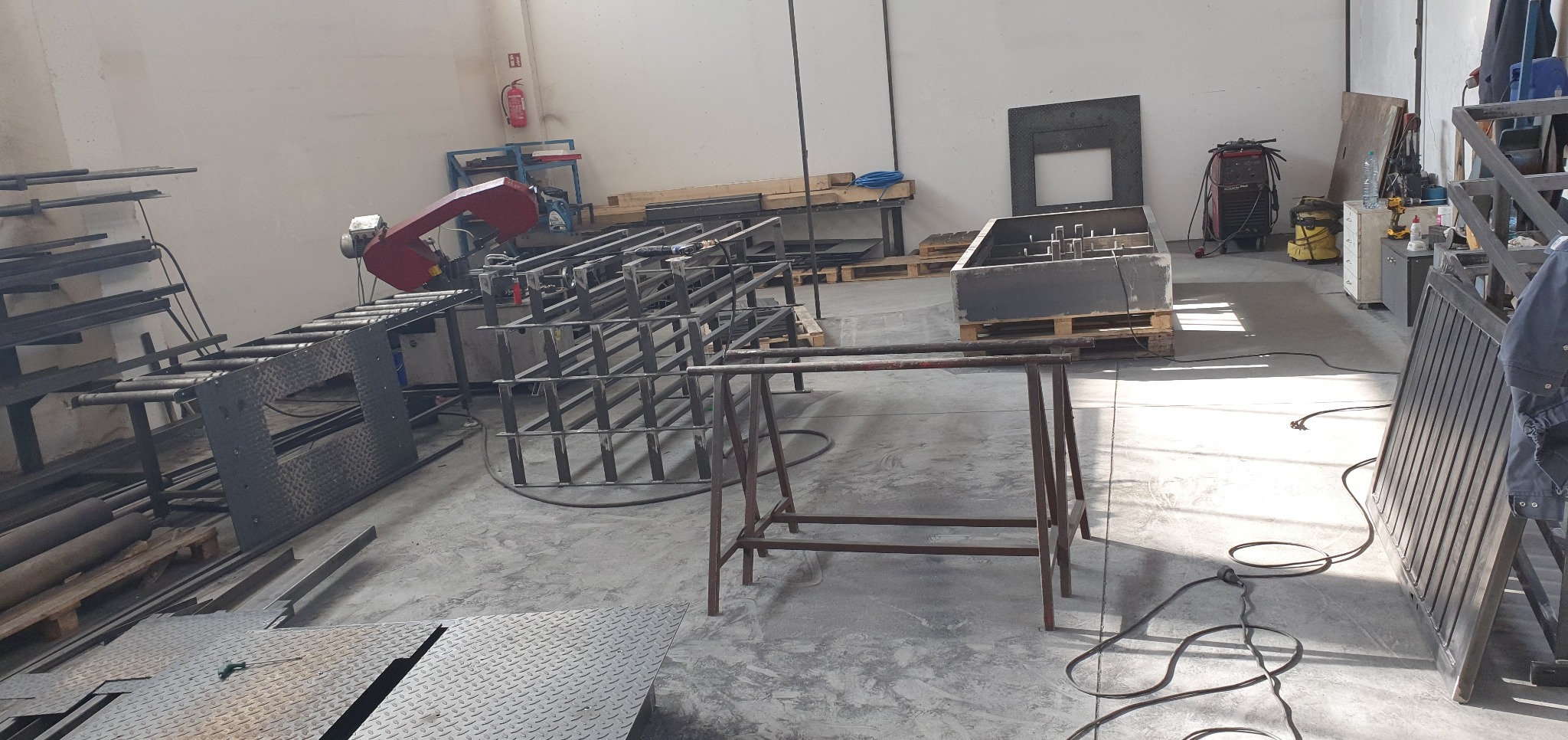 Warsztat z elementami konstrukcji stalowych, w tym kratownice, rama z blachy ryflowanej i metalowa skrzynia na paletach, widoczna piła taśmowa do metalu oraz stanowisko spawalnicze.