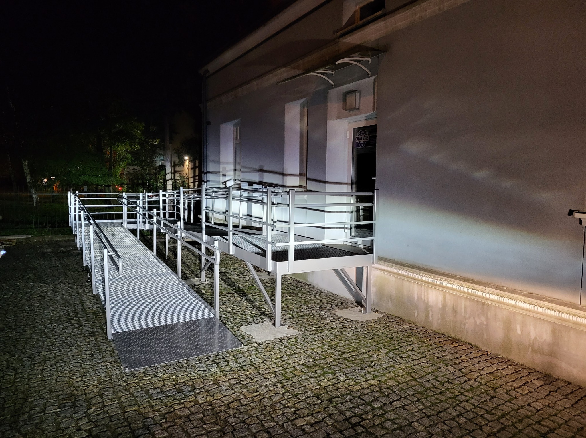 Zewnętrzna rampa dla niepełnosprawnych z metalową konstrukcją i ażurowym pomostem, oświetlona nocą, prowadząca do wejścia budynku z szarą elewacją i szklanym zadaszeniem.