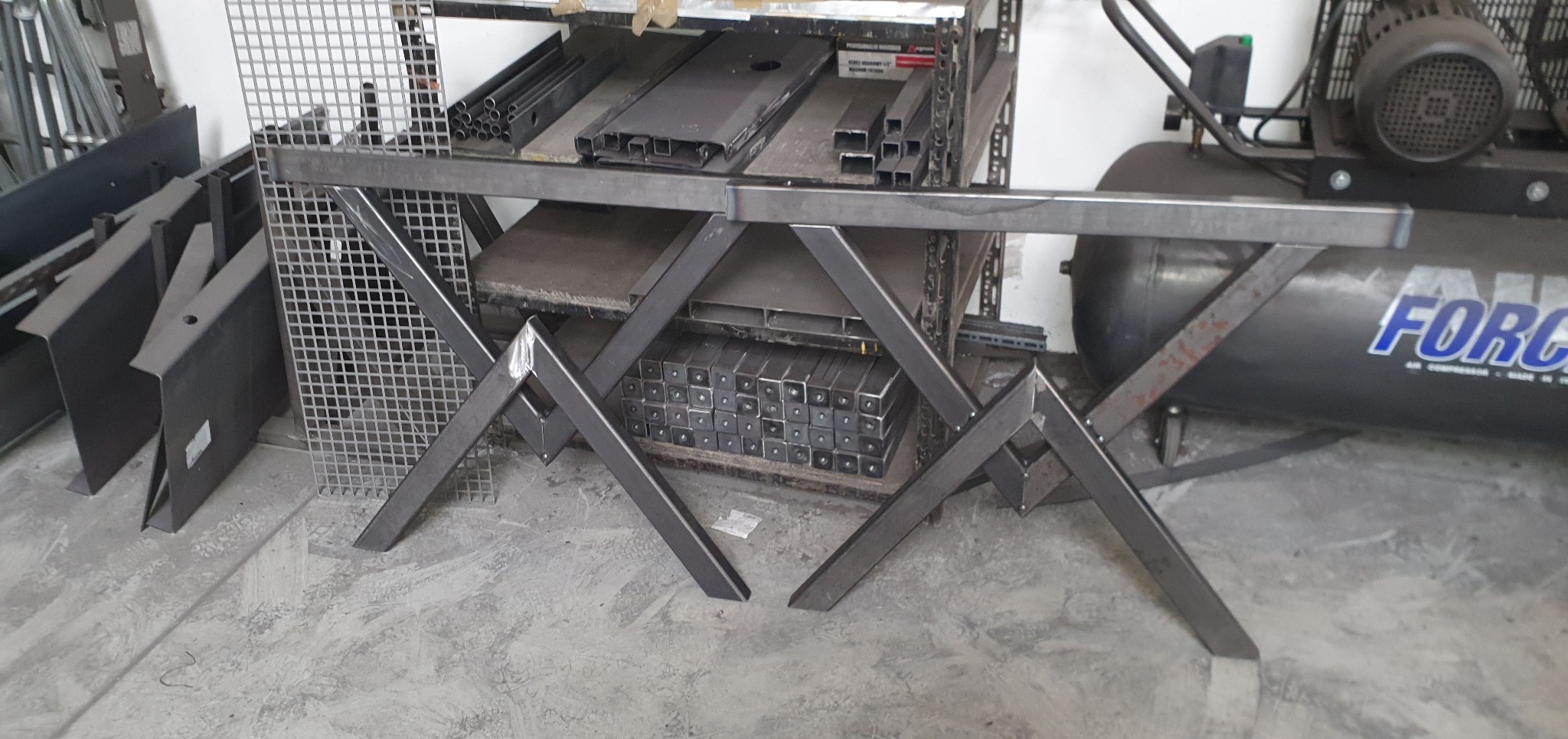 Elementy konstrukcji stalowej w warsztacie: spawana rama stołu z profili, na tle regałów z metalowymi elementami i kompresora.