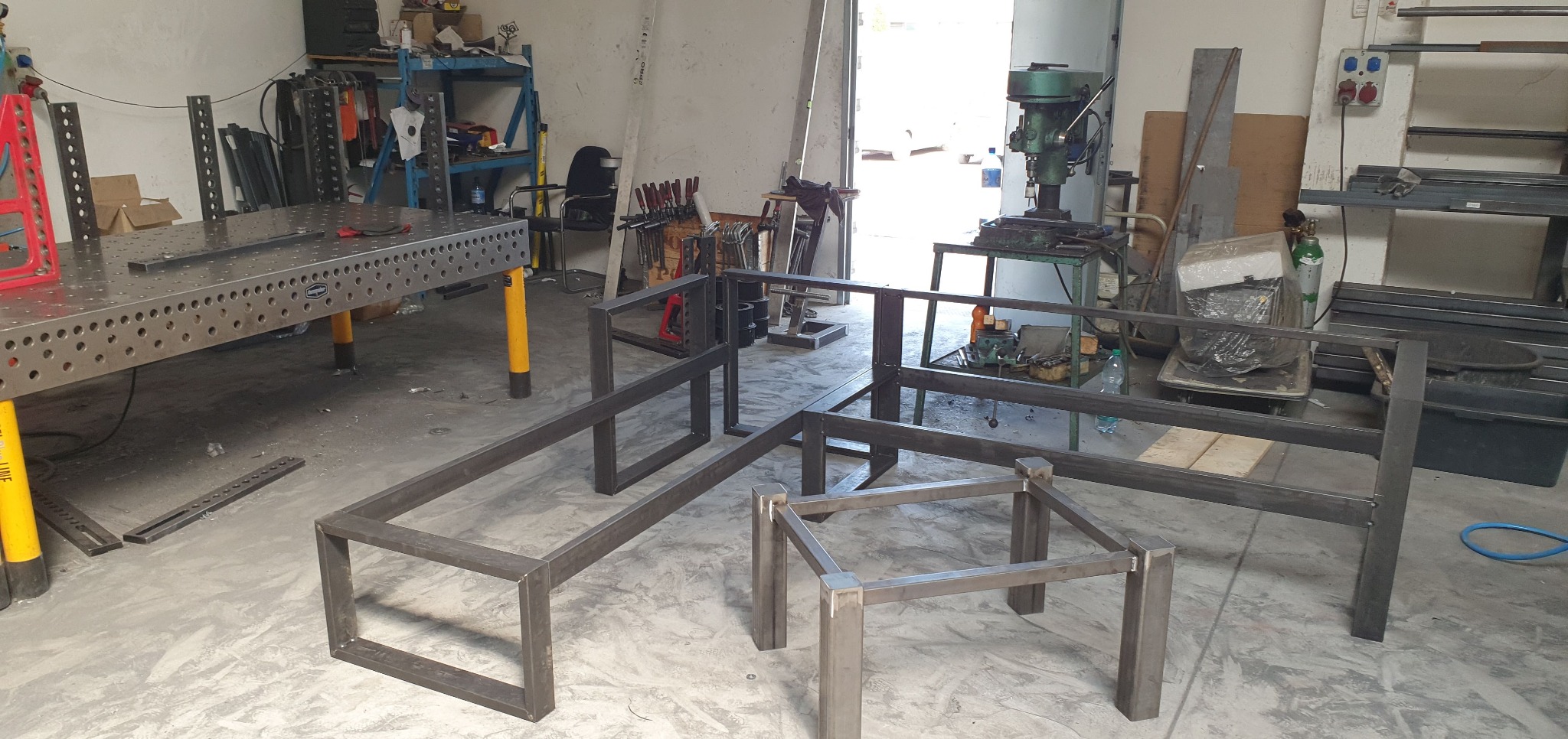 Warsztatowa przestrzeń z metalowymi konstrukcjami w trakcie spawania, widoczny stół spawalniczy z otworami, w tle wiertarka stołowa i narzędzia.