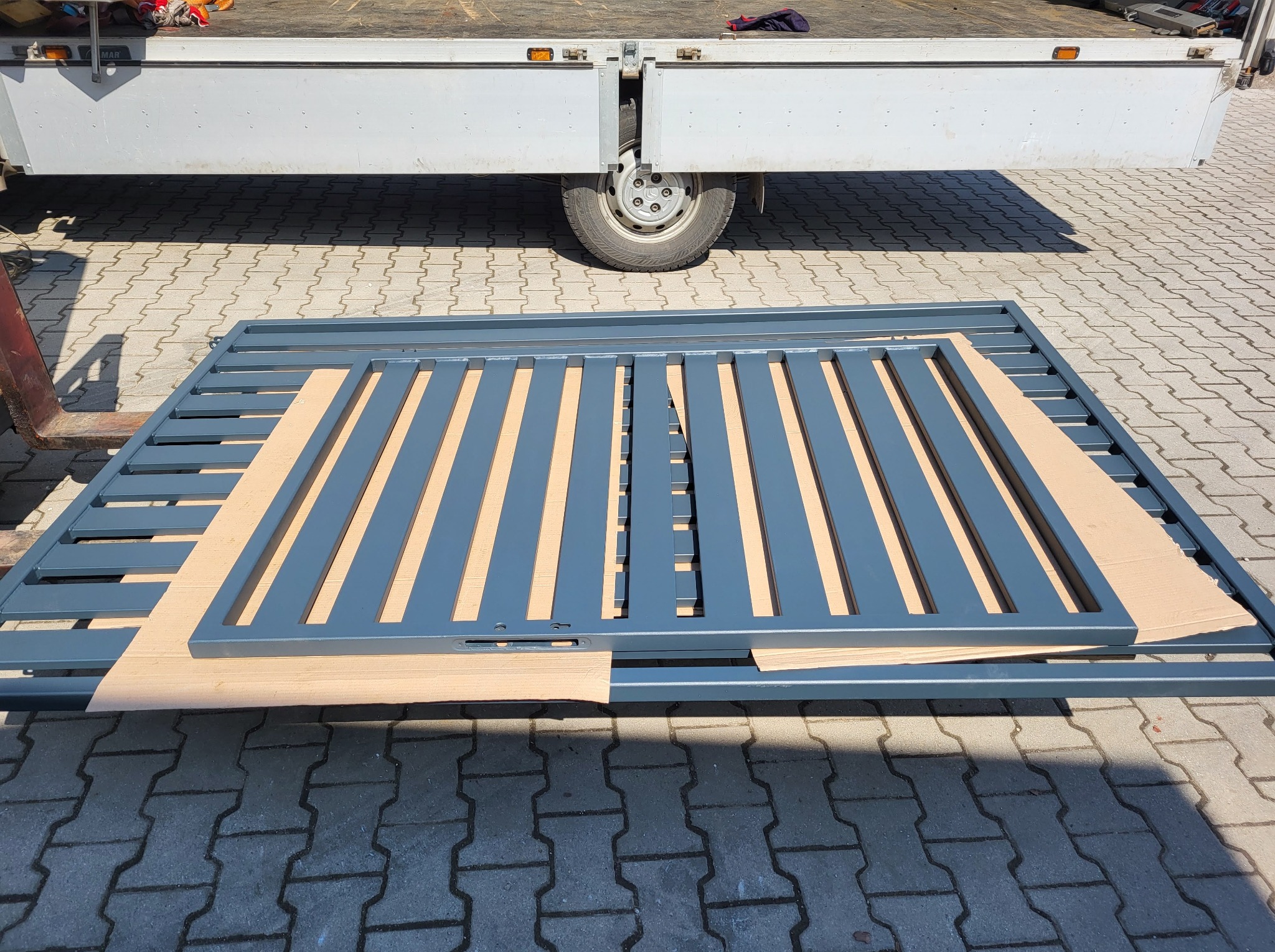 Pomazana na szaro metalowa brama wjazdowa, zabezpieczona kartonem podczas transportu, leży na kostce brukowej, w tle widoczny fragment samochodu dostawczego.