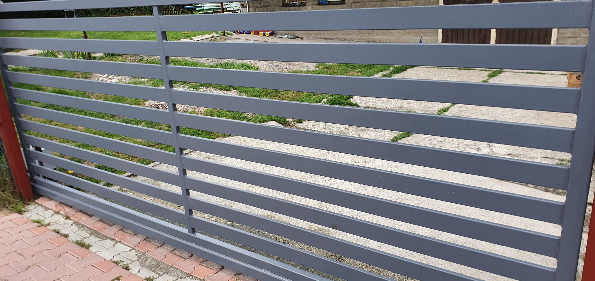 Nowoczesna, pozioma brama wjazdowa z szarych, metalowych profili, widziana z bliska, z kostką brukową u dołu i zielenią w tle.