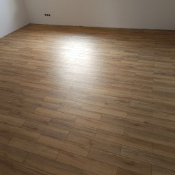 IGO EXPERT SPÓŁKA Z OGRANICZONĄ ODPOWIEDZIALNOŚCIĄ - Wykończona podłoga z paneli imitujących drewno w jasnym pomieszczeniu z białymi ścianami i widocznymi gniazdkami elektrycznymi. Panele ułożone równolegle do ściany, tworząc jednolity wzór.