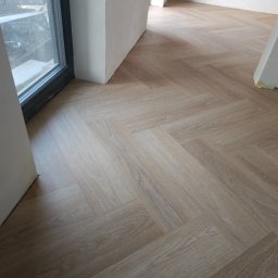 IGO EXPERT SPÓŁKA Z OGRANICZONĄ ODPOWIEDZIALNOŚCIĄ - Jasne panele ułożone w jodełkę, widok z góry, fragment okna i ściany w tle, wykończenie w trakcie prac.