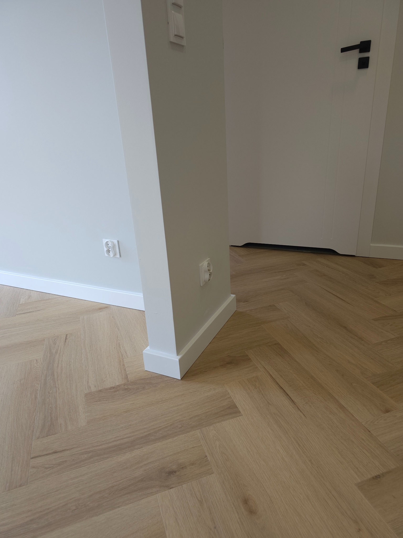 Jasny parkiet ułożony we wzór jodełki francuskiej, białe listwy przypodłogowe i fragment białej ściany z gniazdkiem elektrycznym, w tle białe drzwi z czarną klamką.
