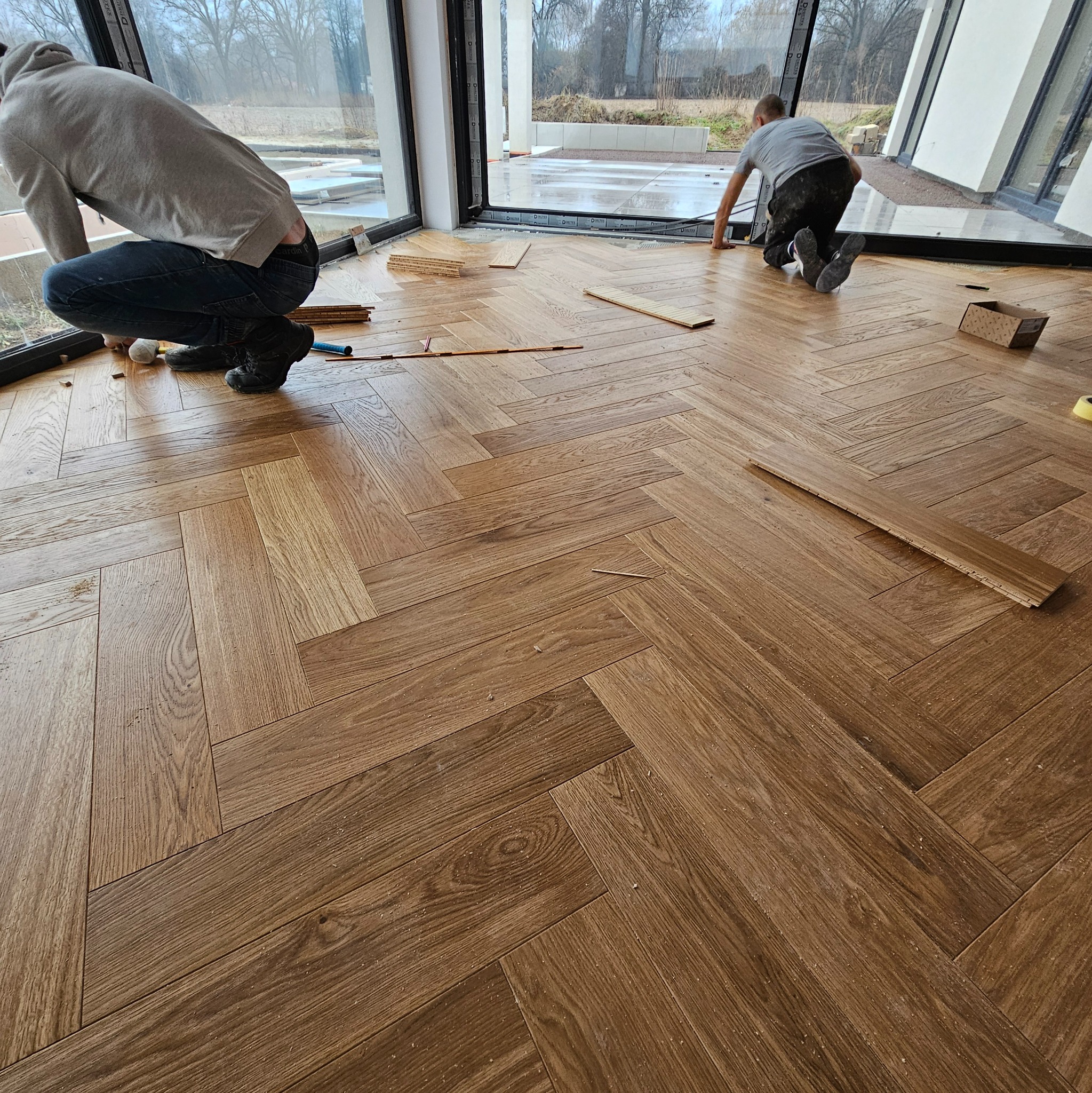 Układanie parkietu w jodełkę w nowoczesnym wnętrzu z dużymi oknami; widoczne narzędzia i fragmenty nieułożonego parkietu.
