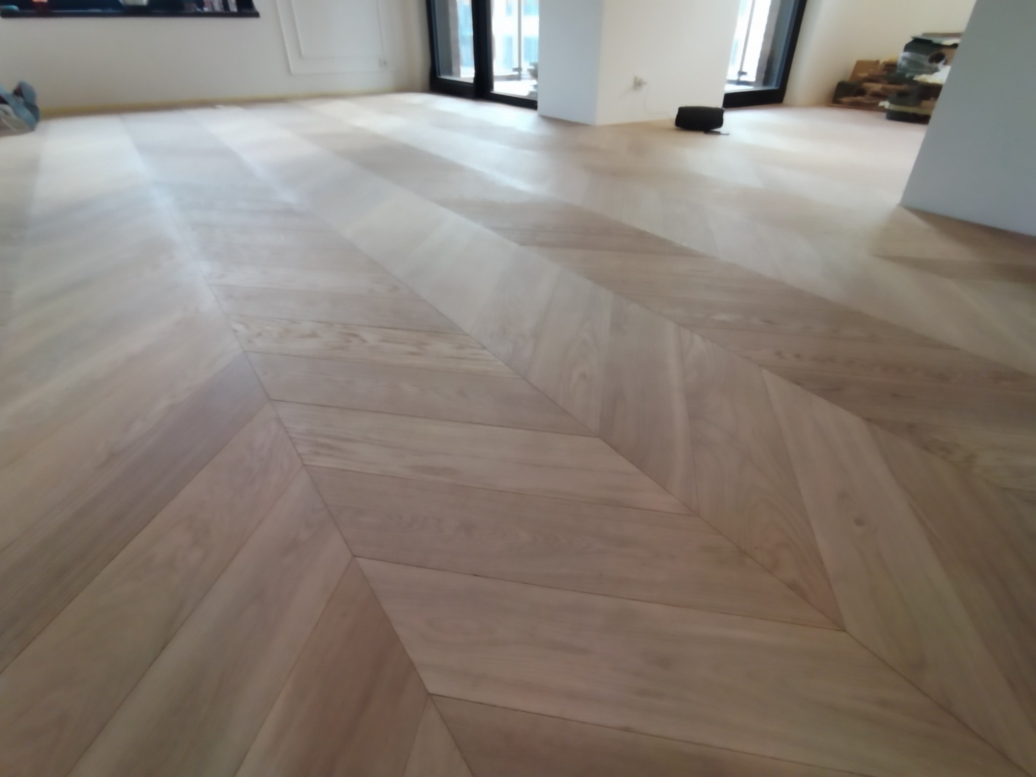 Jasny parkiet ułożony we wzór jodełki francuskiej w przestronnym, jasnym wnętrzu z dużymi oknami.