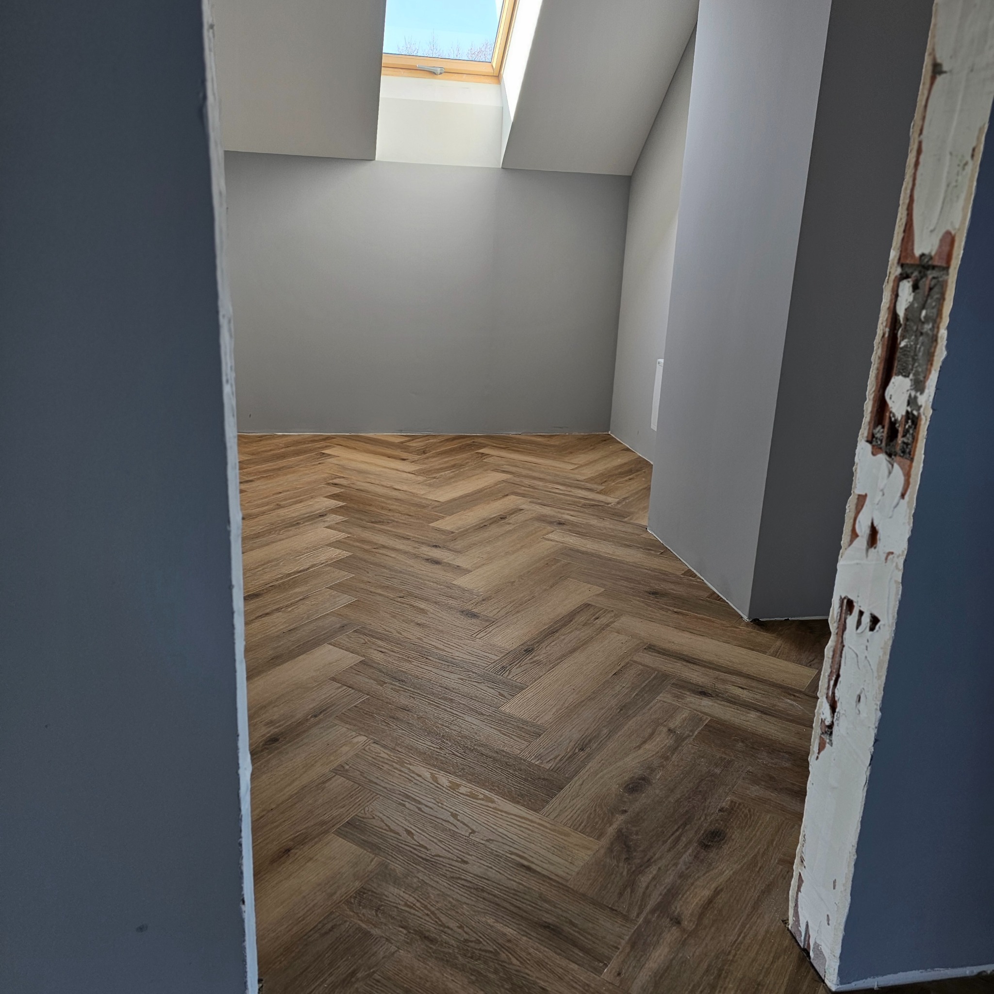 Ukończona podłoga z paneli ułożonych w jodełkę w pokoju z szarymi ścianami i oknem dachowym, widok przez futrynę w trakcie wykańczania.