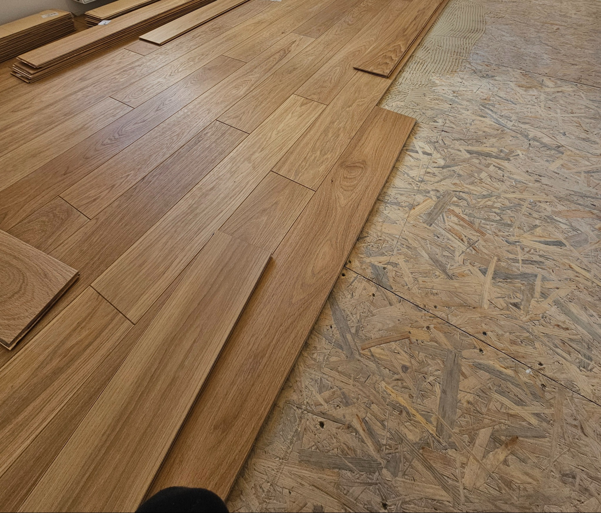 Fragment podłogi w trakcie montażu: ułożone panele o jasnym odcieniu drewna kontrastują z surowym podłożem z płyty OSB, widoczne paczki paneli w tle.