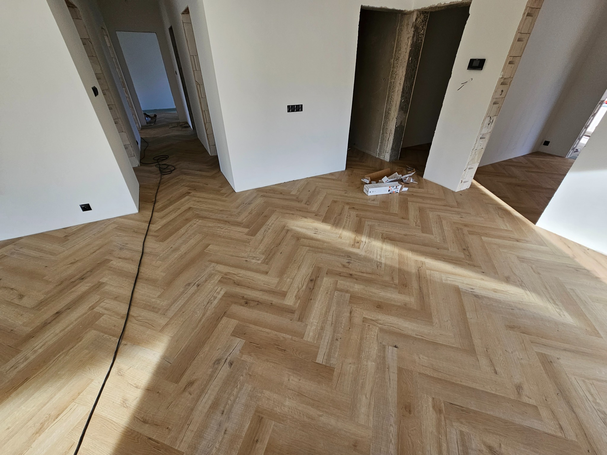 Nowo ułożona podłoga z paneli w jodełkę w jasnym odcieniu drewna, widoczne fragmenty ścian w stanie surowym i kable elektryczne.