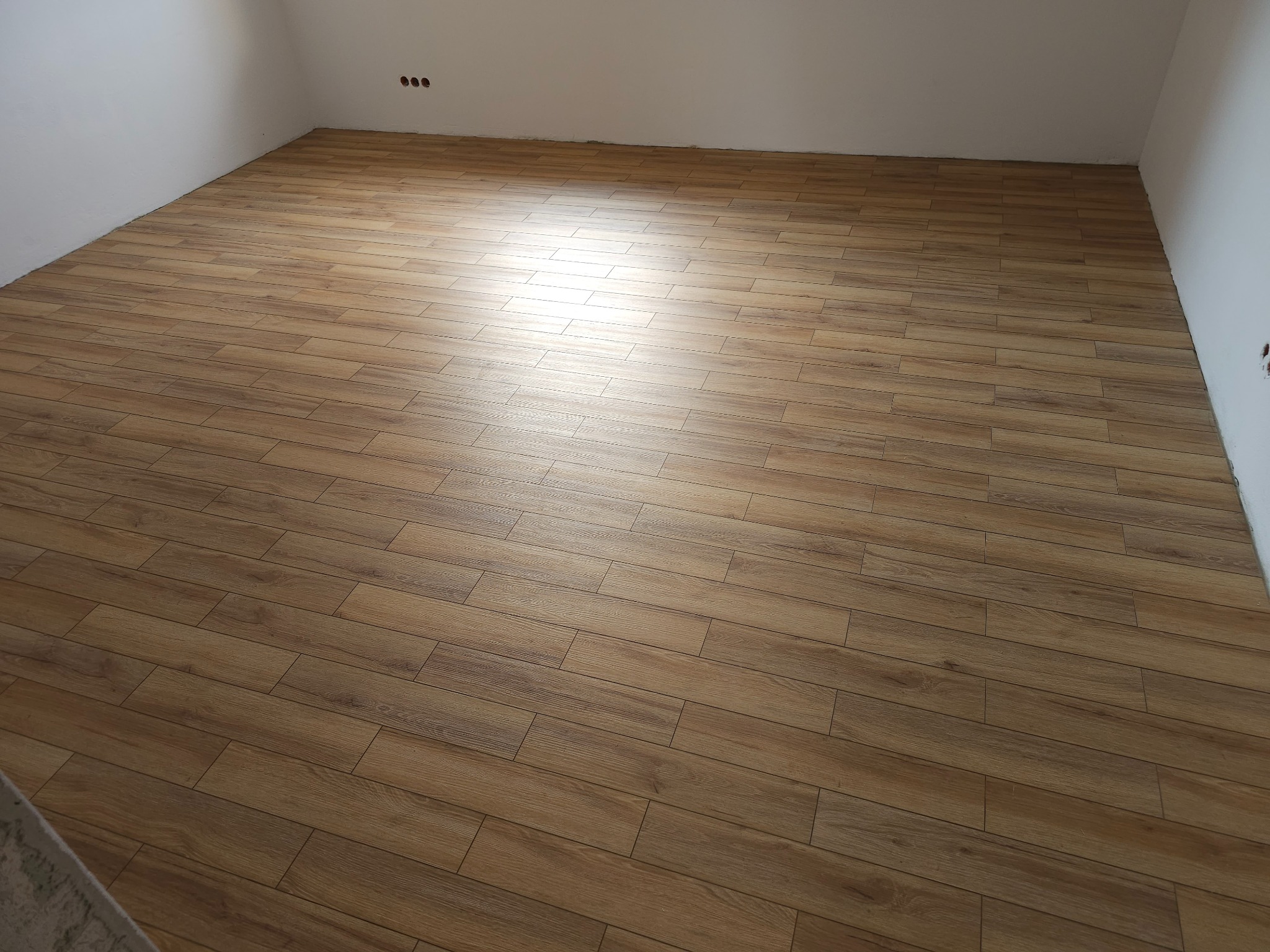 Wykończona podłoga z paneli imitujących drewno w jasnym pomieszczeniu z białymi ścianami i widocznymi gniazdkami elektrycznymi. Panele ułożone równolegle do ściany, tworząc jednolity wzór.