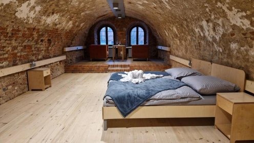 Jasne deski podłogowe w piwnicy z łukowatym ceglanym sklepieniem, na podwyższeniu dwa fotele i stolik, w części sypialnej łóżko z szarą pościelą i niebieskim pledem