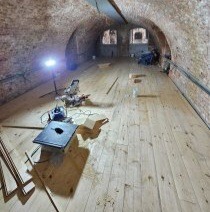 Wnętrze sklepionego pomieszczenia w trakcie remontu, z częściowo ułożoną drewnianą podłogą. Widoczne narzędzia: piła ukośnica, frezarka zagłębiarka, lampa oświetleniowa. W tle okna i ceglany łuk...